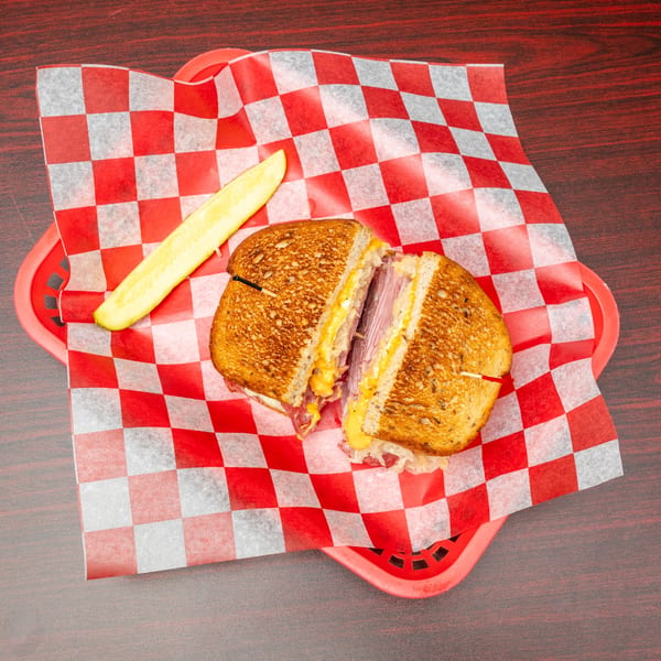 A grilled Reuben sandwich, cut in half, with melted cheese, pastrami, and sauerkraut, served with a pickle spear in a red basket.