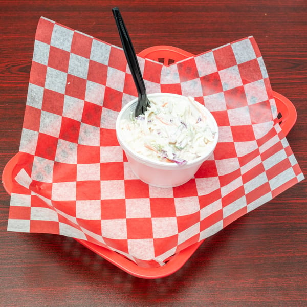 Creamy coleslaw in a white cup with a black fork, resting on red and white checkered paper.