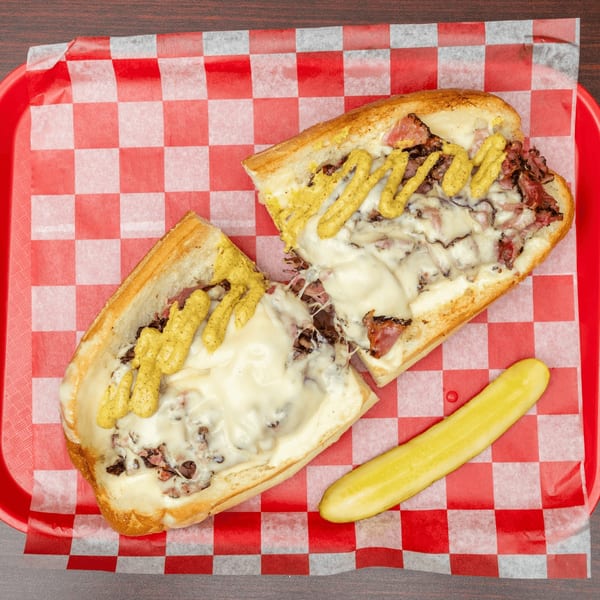 Cheesesteak sandwich with melted cheese and mustard, cut in half, served with a pickle spear.
