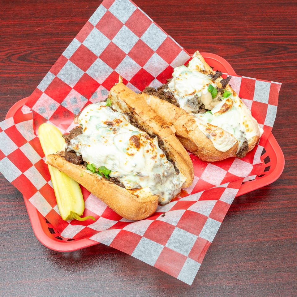 Two cheesy steak and mushroom subs with green peppers and a pickle spear in a red basket.
