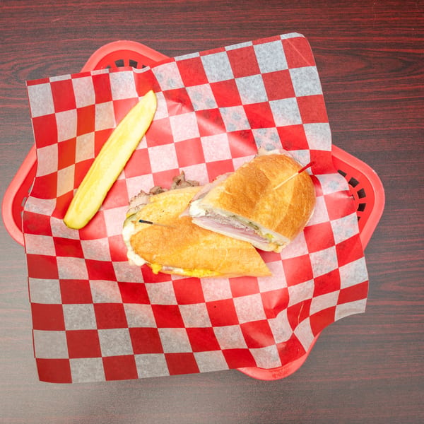 A Cuban sandwich cut in half, served with a pickle spear in a red basket with checkered paper.