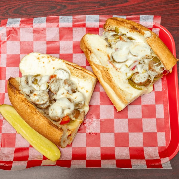 Two sausage and pepper sandwiches with melted cheese and a pickle spear on a red checkered tray.