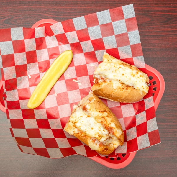 Two halves of a cheesy sub sandwich and a pickle spear in a red basket with checkered paper.