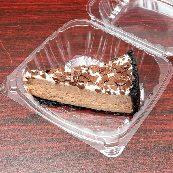 A slice of chocolate cheesecake with whipped cream and chocolate shavings in a clear plastic container.