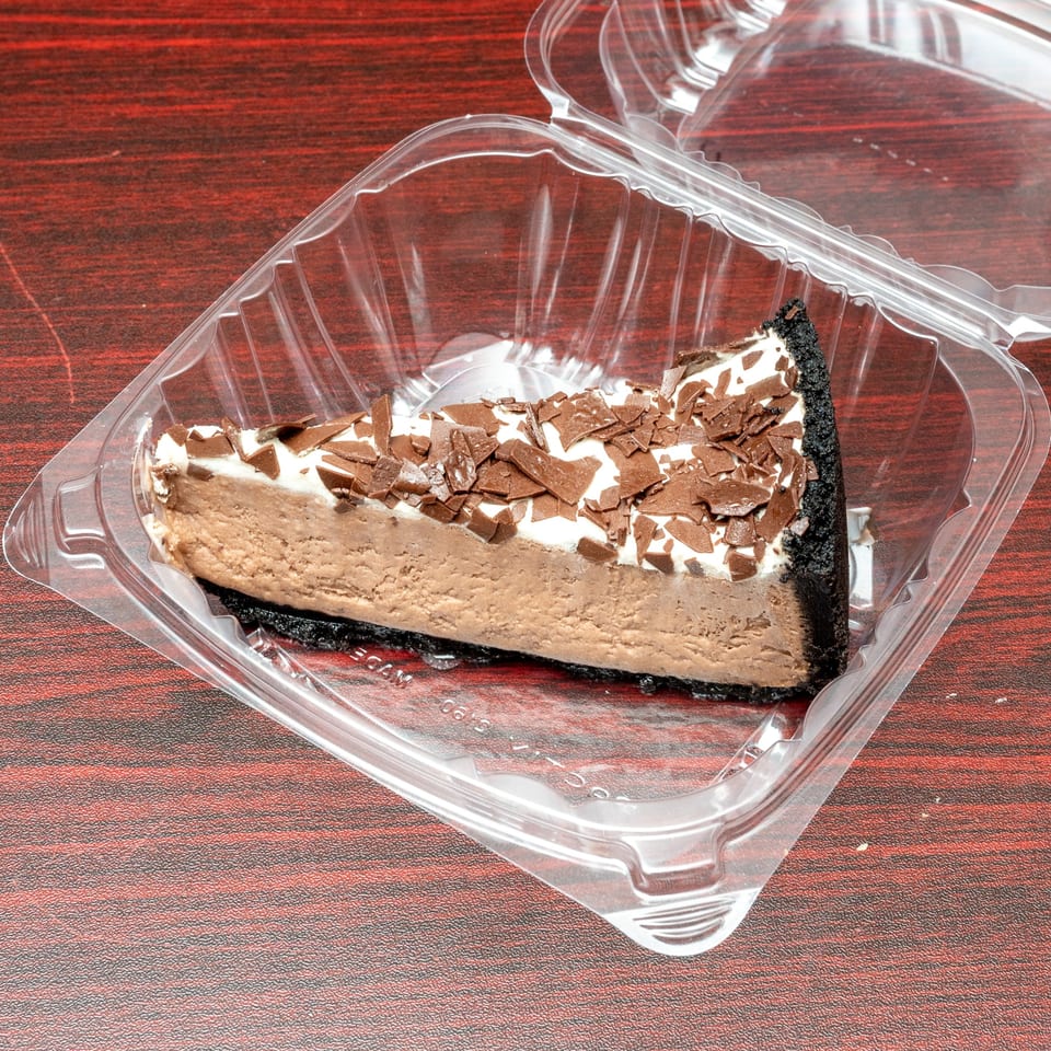 A slice of chocolate cheesecake with whipped cream and chocolate shavings in a clear plastic container.